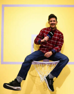 Chico con camisa de cuadros rojos y vaqueros está sentado sobre un aro de baloncesto lila y está sujetando un micrófono de fibwi.
